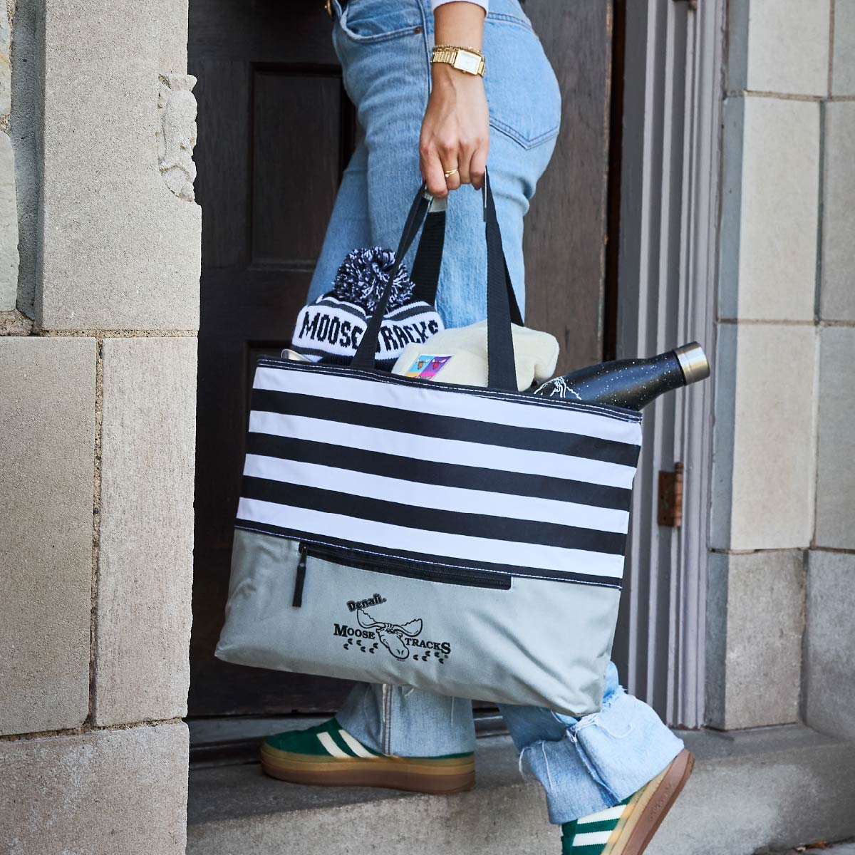 A person walking in through a door while holding the Moose Tracks Cooler Bag. The bag has a striped design, zipper and Moose Tracks logo on the front.