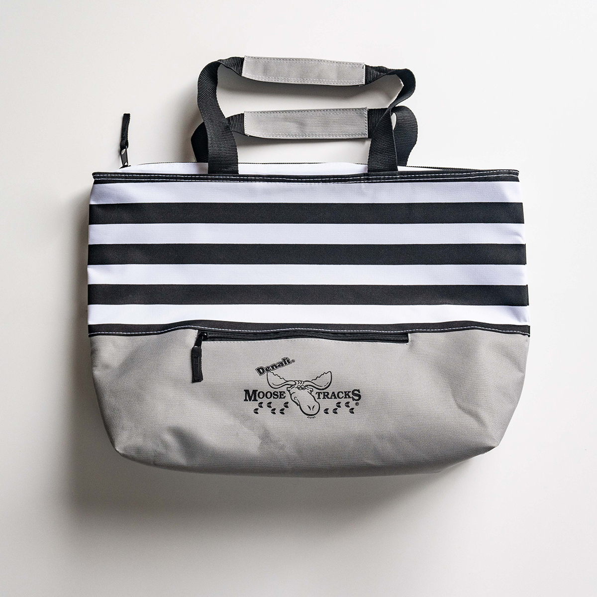 The Moose Tracks Cooler Bag laying on a flat surface with the front showing. The bag has a striped design and features a zipper and Moose Tracks logo on the front.