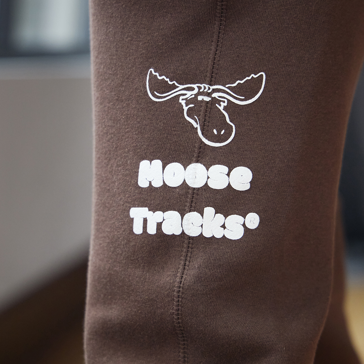 A close-up picture of vanilla-colored screen printing on brown sweatpants, which reads, "Moose Tracks," and a moose icon