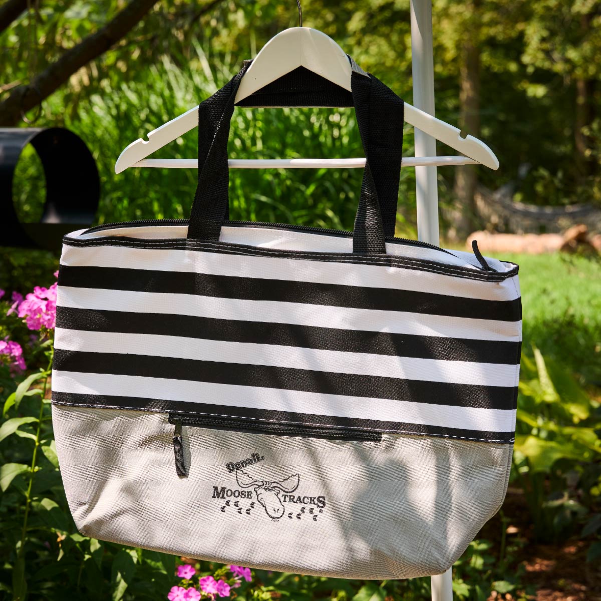 The Moose Tracks Cooler Bag hanging on a hanger outside by a garden.