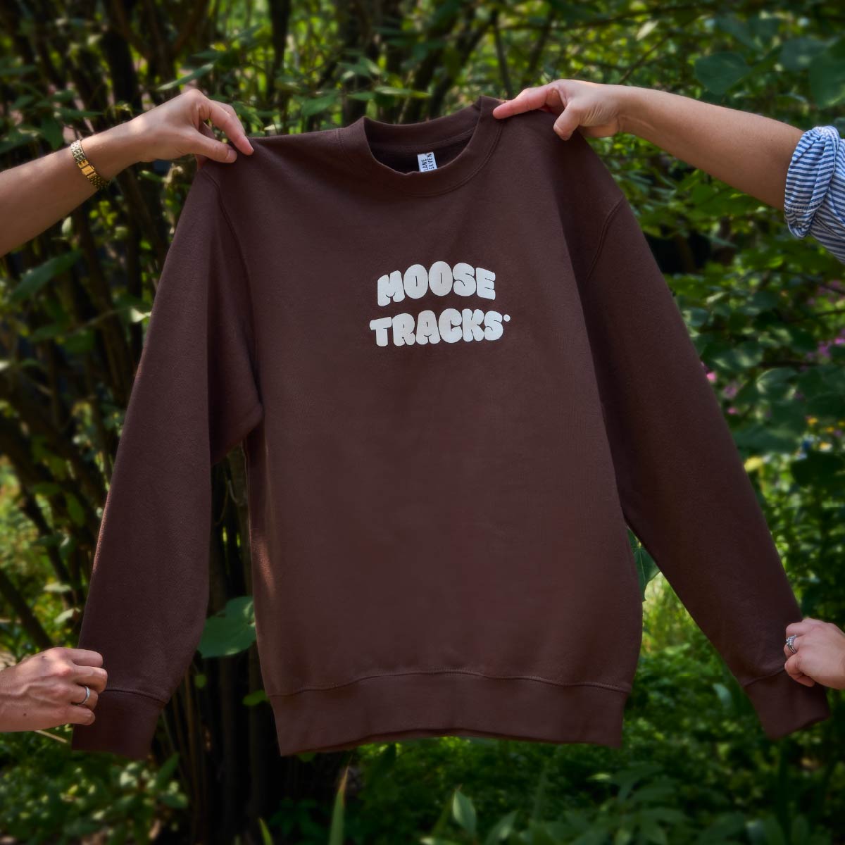 Two sets of hands holding a brown crewneck sweatshirt that reads, "Moose Tracks" in vanilla-colored screen printing.
