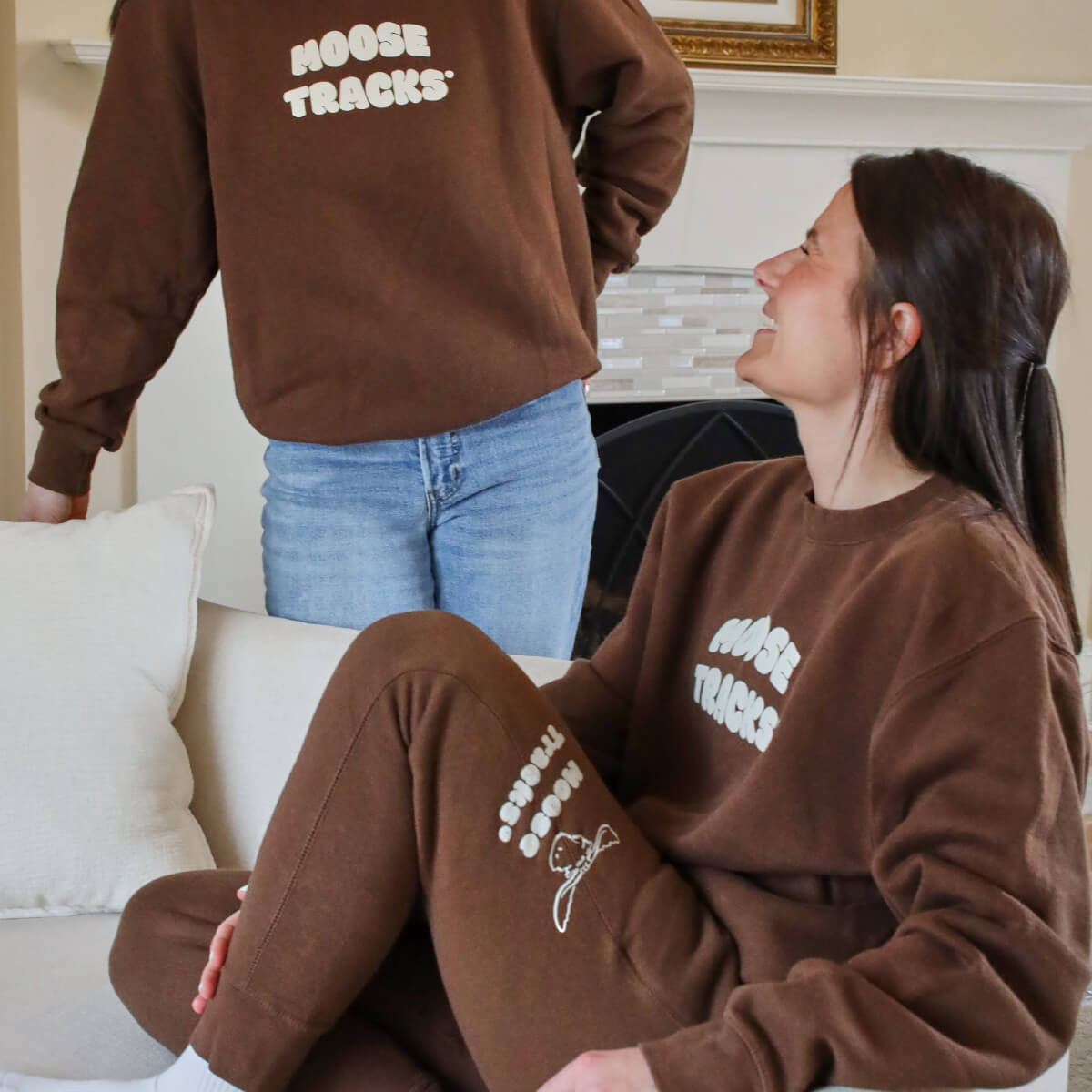 Two women are wearing matching brown sweatshirts with white text reading "MOOSE TRACKS" on the front. One woman is standing and the other is seated and looking up. The seated woman is wearing brown sweatpants that have a white moose head logo and "MOOSE TRACKS" printed on the side.