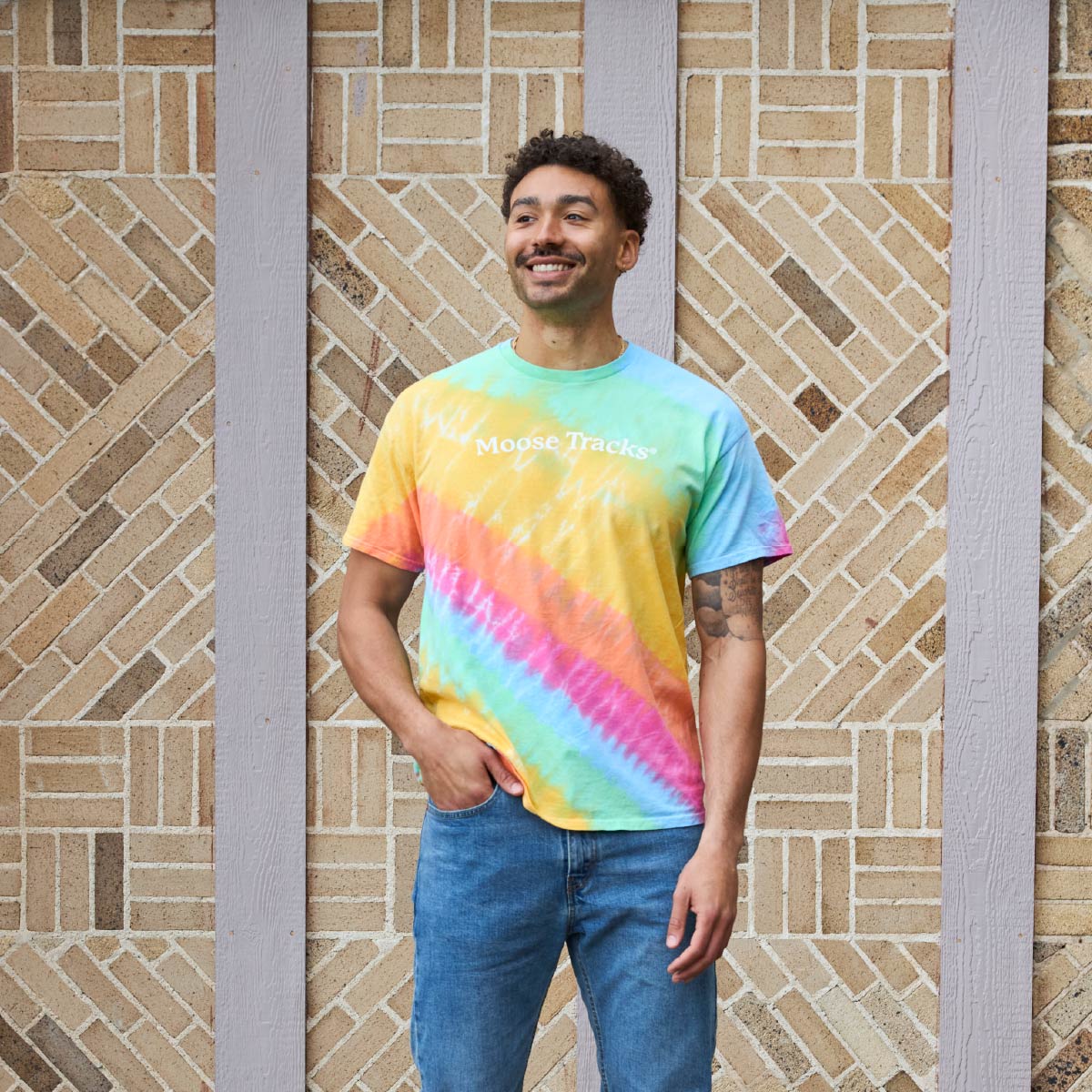 A person wearing the Moose Tracks Tie-Dye T-Shirt. The shirt features a rainbow pastel tie-dye pattern with white screen-printed text in the center that reads "Moose Tracks".