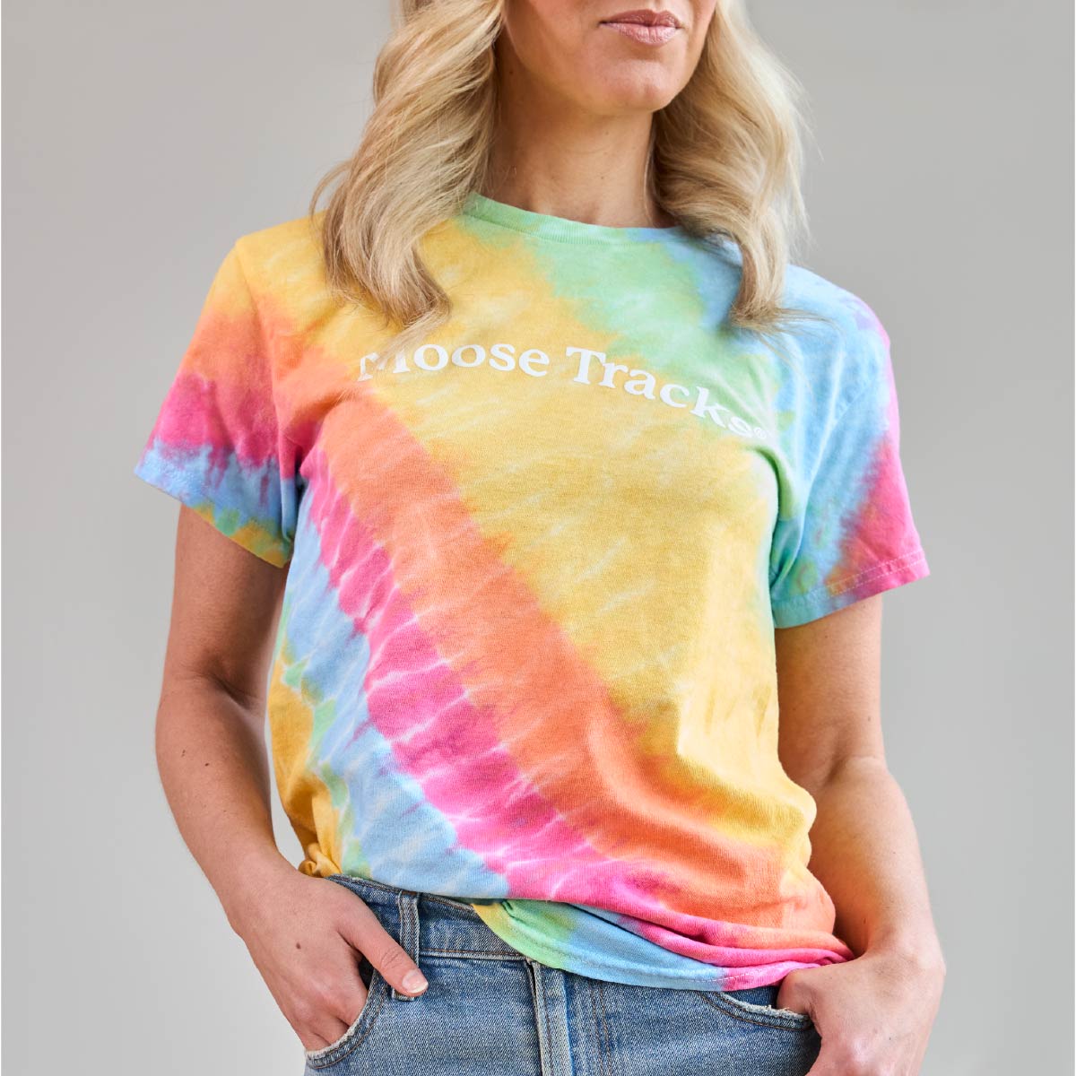 A closer view of someone wearing the Moose Tracks Tie-Dye T-Shirt. The shirt features a rainbow pastel tie-dye pattern with white screen-printed text in the center that reads "Moose Tracks".