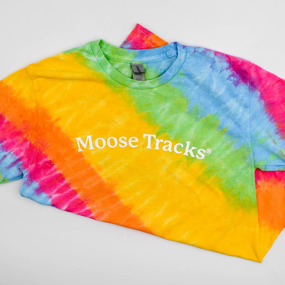 An aerial view of the Moose Tracks Tie-Dye T-Shirt with the bottom folded back, laying on a flat surface. The shirt features a rainbow pastel tie-dye pattern with white screen-printed text in the center that reads "Moose Tracks".