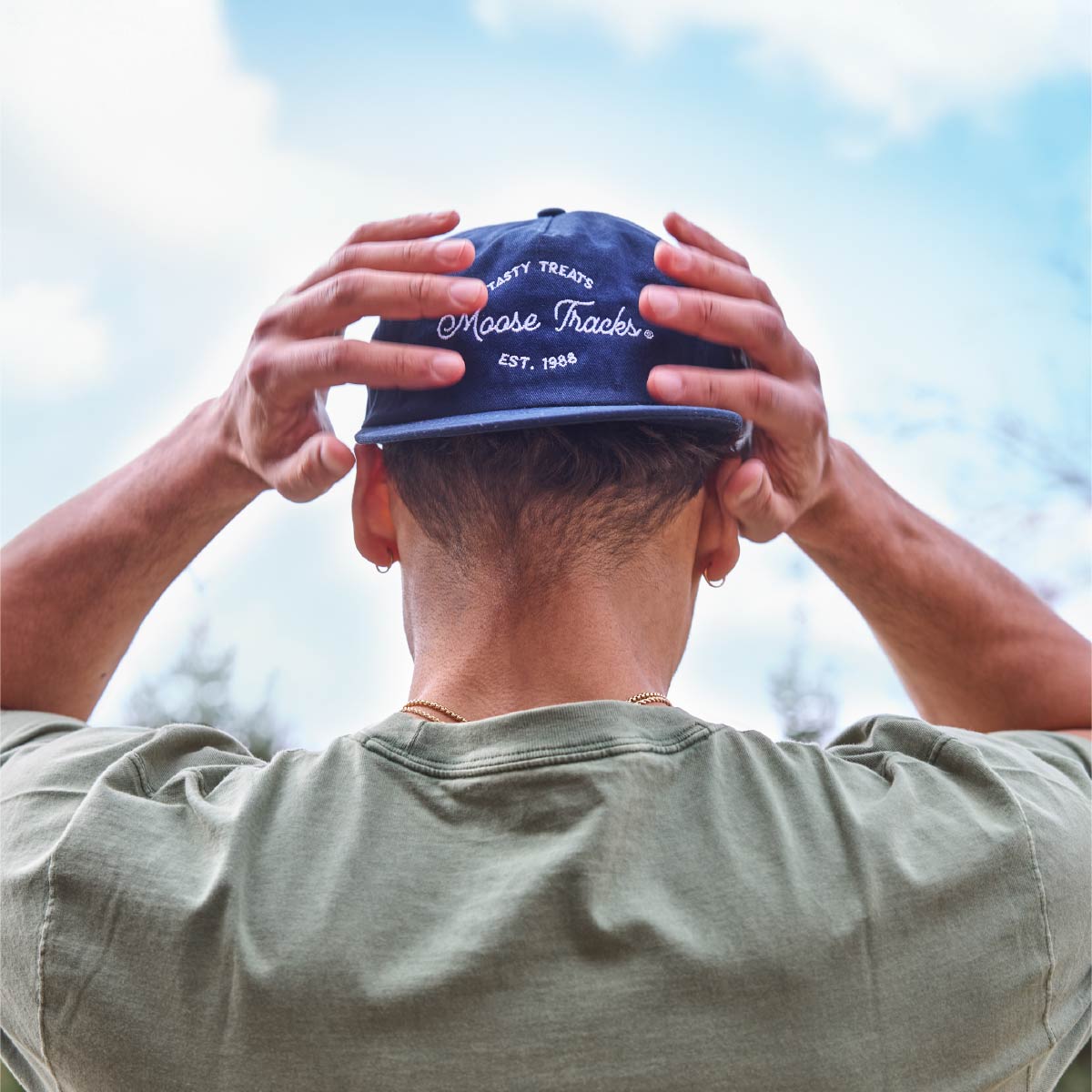 A person wearing the Moose Tracks Navy Snapback Hat backwards with their hands on the hat. The hat is navy with white-colored embroidered text reading "Tasty Treats Moose Tracks EST. 1988".