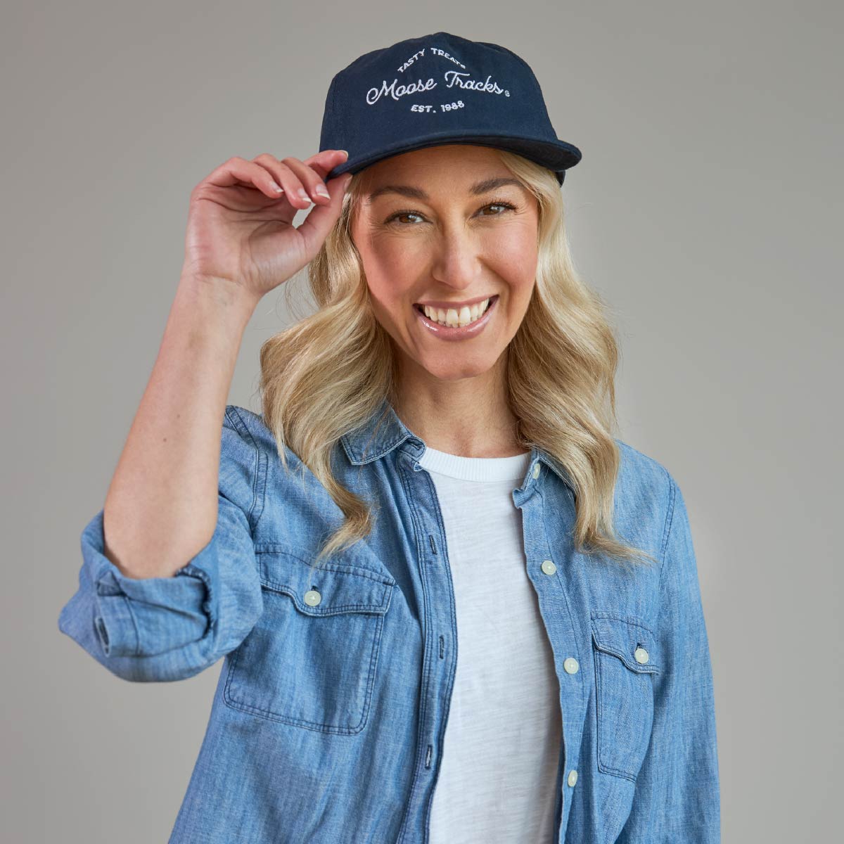 A smiling person wearing the Moose Tracks Navy Snapback Hat with their hand on the tip of the hat. The hat is navy with white-colored embroidered text reading "Tasty Treats Moose Tracks EST. 1988".