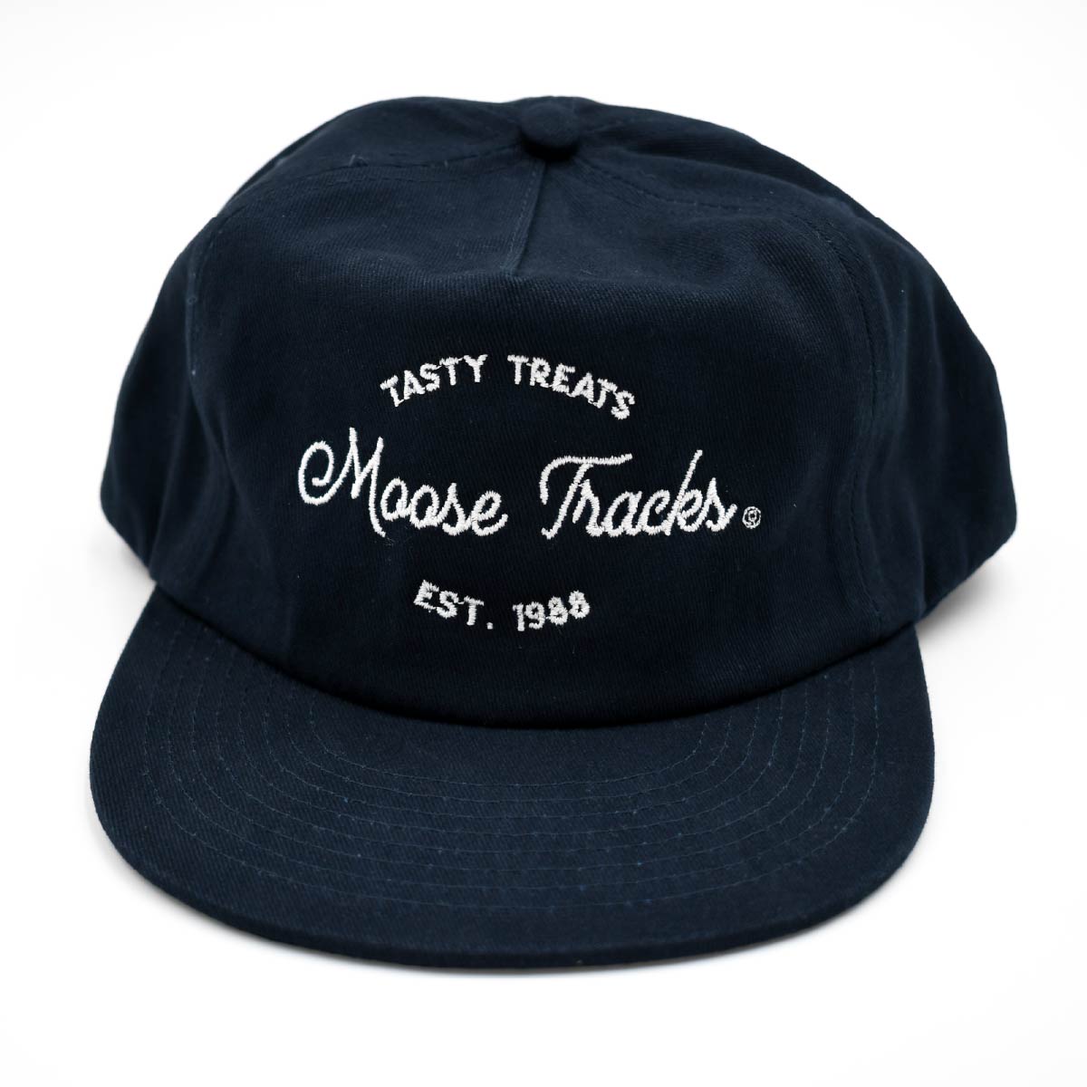 A front view of the Moose Tracks Navy Snapback Hat placed on a flat surface with a white background. The hat is navy with white-colored embroidered text reading "Tasty Treats Moose Tracks EST. 1988".