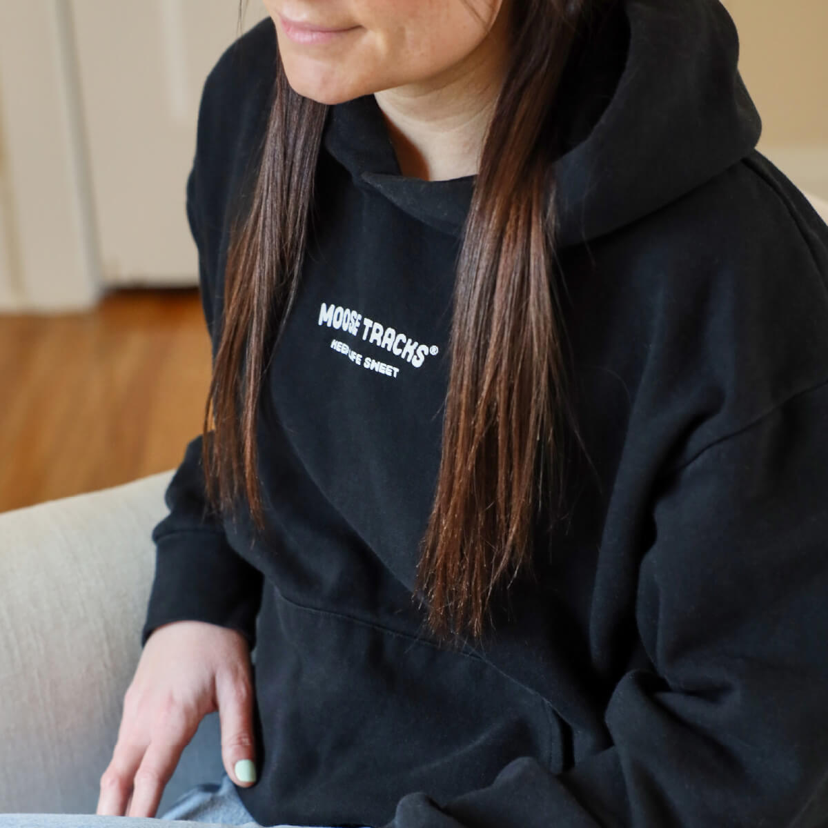 A woman with long brown hair sitting on a sofa and wearing a black hoodie that reads, "Moose Tracks Keep Life Sweet" across the chest in white letters.