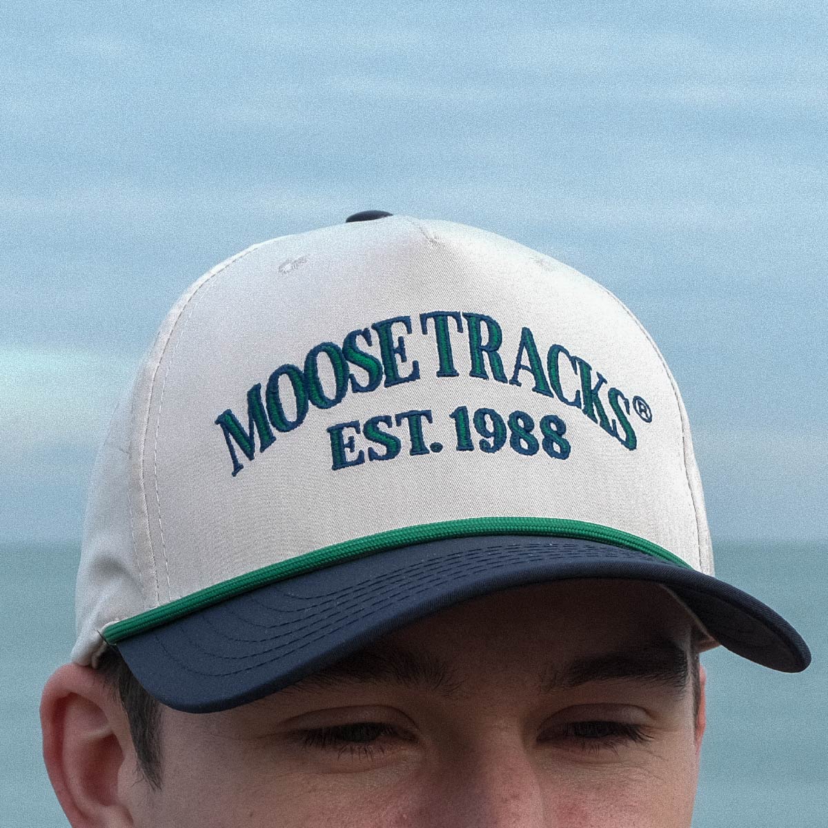 A man wearing a Moose Tracks two-tone snapback rope hat outdoors. The hat features a light tan crown, navy bill, and green rope across the bill with "Moose Tracks EST. 1988" printed on the front in green letters with a navy outline.