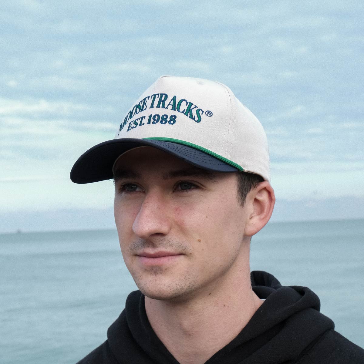 A man wearing a Moose Tracks two-tone snapback rope hat outdoors. The hat features a light tan crown, navy bill and green rope across the bill, and has "Moose Tracks EST. 1988" printed in green letters with a navy outline.