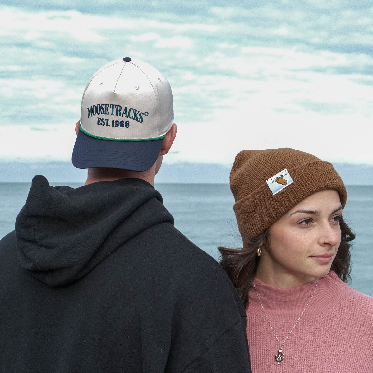 A man and woman standing next to each other wearing a Moose Tracks two-tone snapback rope hat and brown Moose Tracks rib-knit cuffed beanie, surrounded by water and clouds in the sky.