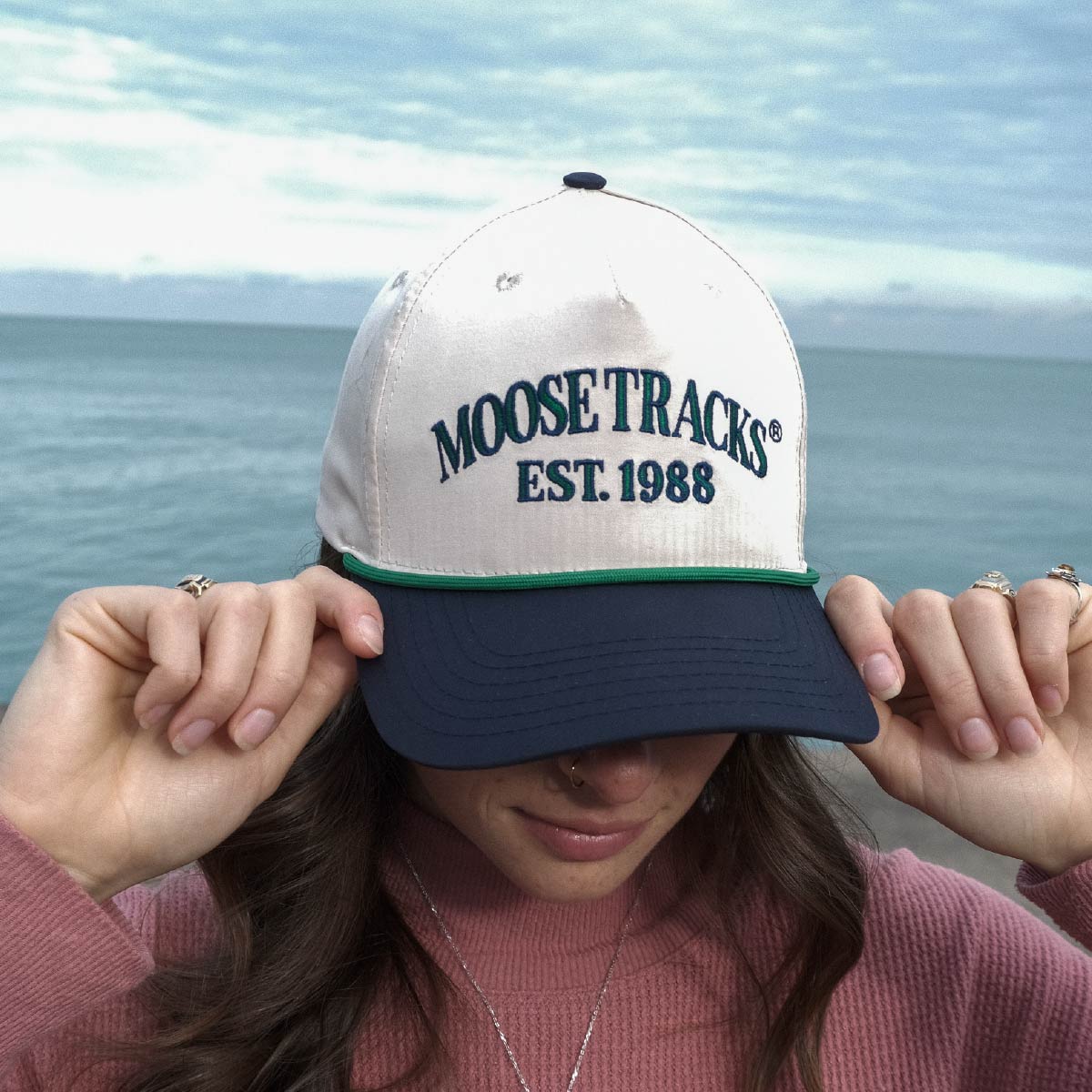 A woman wearing a Moose Tracks two-tone snapback rope hat, slightly facing downwards with their hands on the bill. The hat is primarily light tan with a navy bill, has a green rope across the bill and "Moose Tracks EST. 1988" printed on the front in green letters with a navy outline.