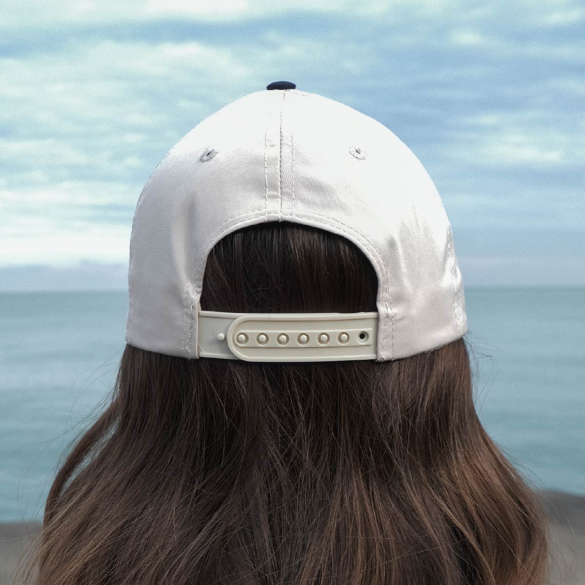 A woman wearing a Moose Tracks two-tone snapback rope hat stands outwards, against a cloudy sky and lakefront. The hat features a light tan crown, light tan clasp and navy button showing on the top.