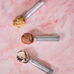 Three aluminum ice cream scoops laying on a marble pink surface, each with a scoop of ice cream in them. The handles are laser-engraved with text that reads, "This is my Moose Tracks scoop".