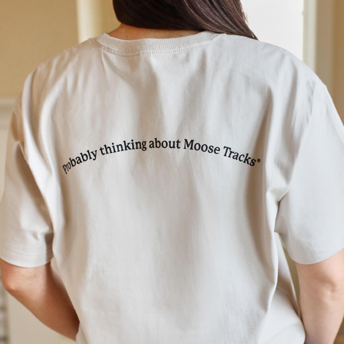A woman wearing a light-colored t-shirt with text that reads, "Probably thinking about Moose Tracks" on the back.