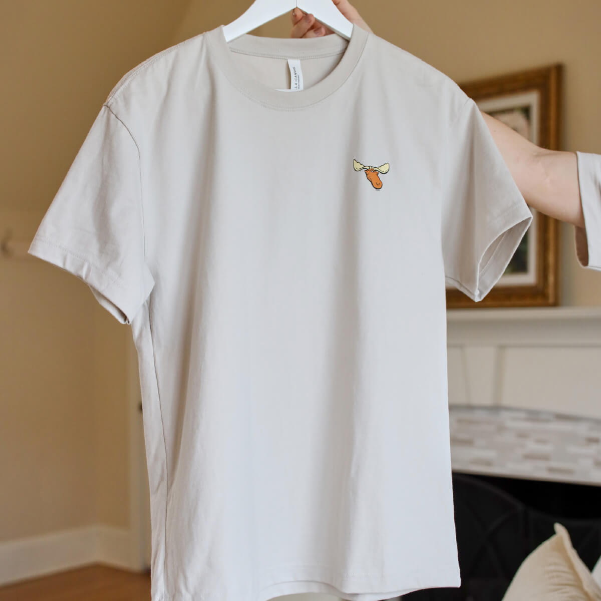 A light-colored t-shirt hanging on a hanger, featuring a moose head logo on the left chest.